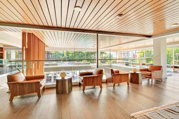 An upper-level view of a clubhouse sky lounge with modern wood armchairs, side tables, and glass railings overlooking large windows and the floor below.