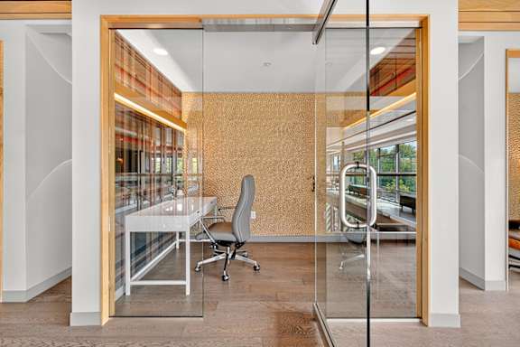 A glass-enclosed private workspace with a white desk, gray office chair, and accent wall in a modern clubhouse setting.