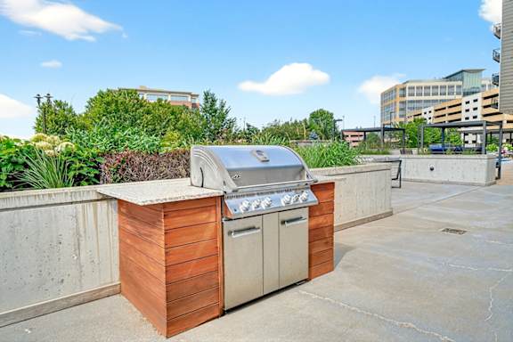 A built-in stainless steel grill with a wood and concrete base set on a spacious outdoor lounge patio surrounded by greenery.