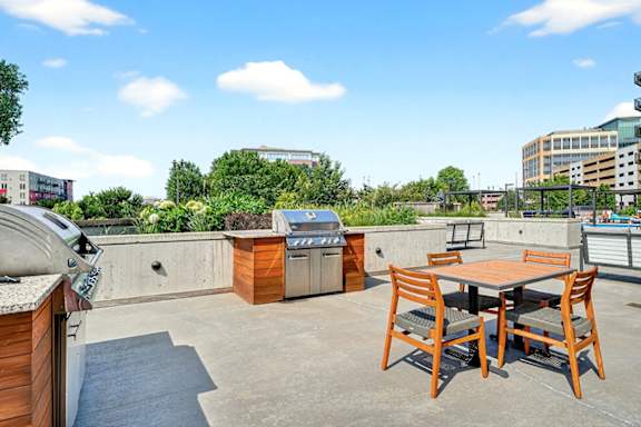An outdoor lounge with built-in stainless steel grills, a wooden dining table with four chairs, and surrounding greenery and cityscape.