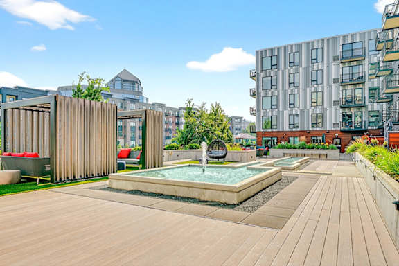 An open courtyard with a rectangular water fountain, covered lounge seating areas with red cushions, an egg chair, and surrounding modern apartment buildings.