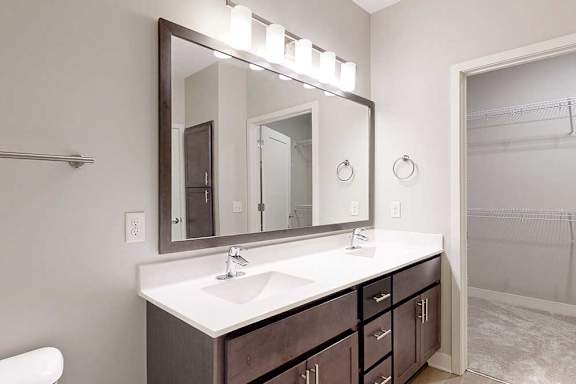 A bathroom with a double sink marble vanity, oversized mirror, dark wooden cabinets, and a door open to walk-in closet.