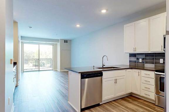 A kitchen with white wooden cabinets and stainless steel appliances, open to a living area with hardwood-style flooring.