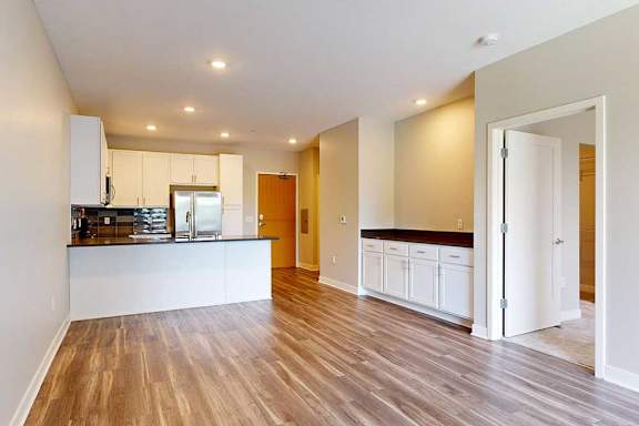 A living room with hardwood-style flooring and kitchen with white wooden cabinets.