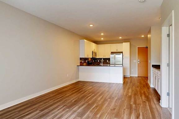 A living room with hardwood-style flooring and kitchen with white wooden cabinets.