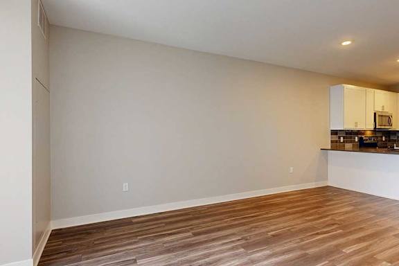 A living room with hardwood-style flooring and kitchen with white wooden cabinets.