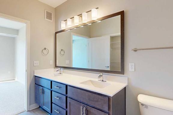 A bathroom with a double sink marble vanity, oversized mirror, dark wooden cabinets, and a door open to a bedroom.