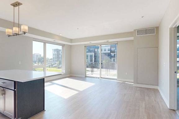 A kitchen with dark wood cabinets and a large island, adjacent to a living room with floor-to-ceiling windows and a sliding glass door to the balcony.