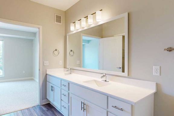 A bathroom with a double sink marble vanity, oversized mirror, white wooden cabinets, and a door open to a bedroom.