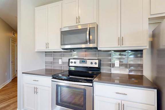 A close up on a stainless steel range and microwave in a kitchen with white wooden cabinets, quartz countertops, and a designer tile backsplash.