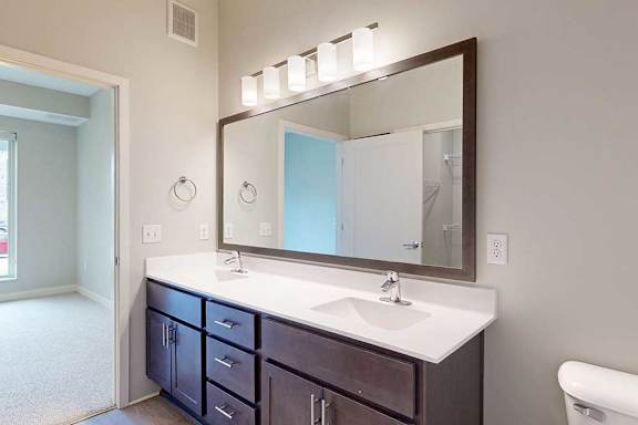 A bathroom with a double sink marble vanity, oversized mirror, dark wooden cabinets, and a door open to a bedroom.