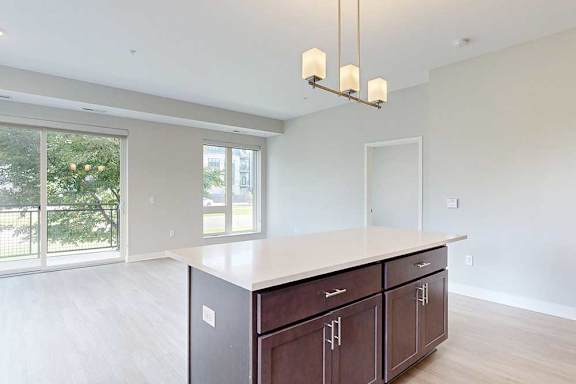 A kitchen with dark wooden cabinets and a large island, in front of a spacious living room.