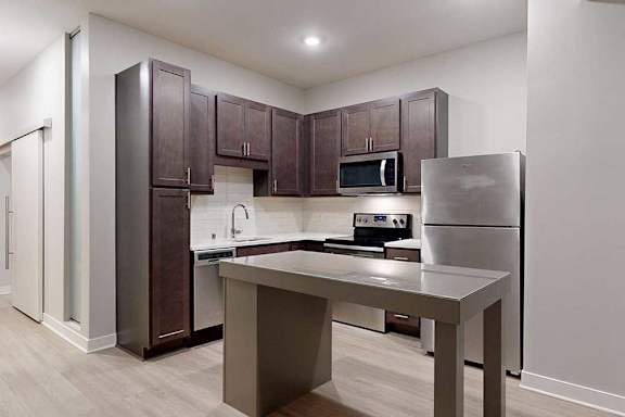 A kitchen with dark wood cabinets, quartz countertops, an island, stainless steel appliances, and a designer backsplash.