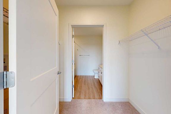 Open white door leads to a carpeted walk-in closet with wire shelving, opening into a bathroom with wooden floor and visible sink and toilet.