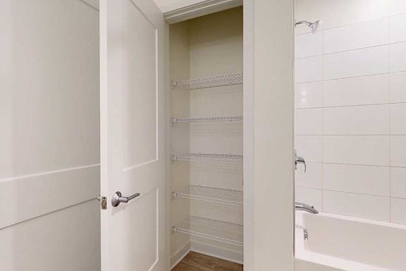 Open door reveals a linen closet with white wire shelves beside a bathtub featuring white tiled walls
