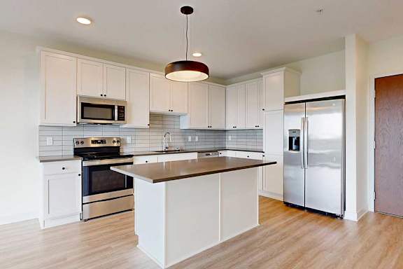 Modern kitchen with sleek white cabinets, stainless steel appliances, and light wood flooring. Central island with a black countertop under a stylish pendant light.