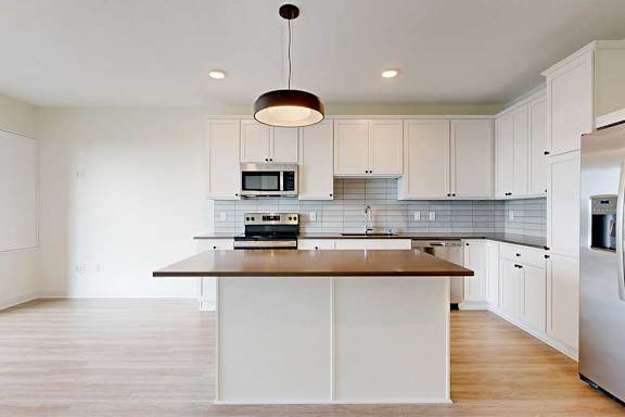 Modern kitchen with sleek white cabinets, stainless steel appliances, and light wood flooring. Central island with a black countertop under a stylish pendant light.