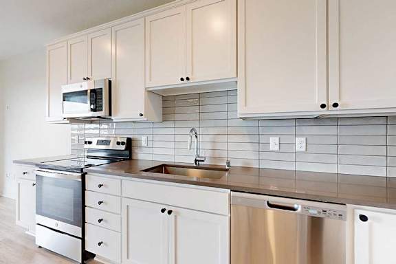 Modern kitchen with white cabinets, stainless steel appliances, and a sleek gray tile backsplash. Includes a stove, microwave, sink, and dishwasher.