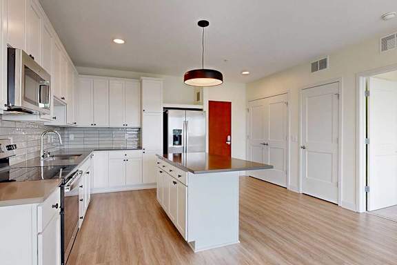 Modern kitchen with white cabinets, stainless steel appliances, and a central island under a dark pendant light. Light wood floor adds warmth.