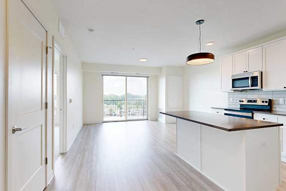 Bright, modern kitchen and living area with wood floors, white cabinets, a black chandelier, and a view through large sliding glass doors to a balcony.