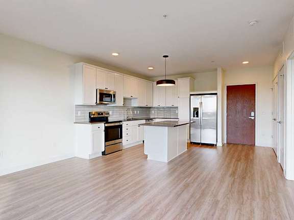 Modern kitchen with white cabinets, stainless steel appliances, wooden floor, and a central island under a sleek pendant light, creating a clean, spacious feel.