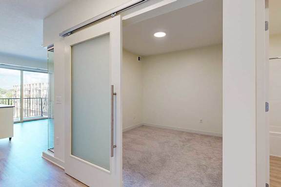 A bright room features a frosted glass sliding door, beige carpeting, and modern wood flooring. A large window offers a view of a balcony and buildings.