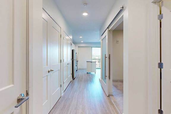 Modern, bright apartment hallway with wood flooring, white walls, and multiple doors. An open kitchen with stainless steel appliances is visible ahead.