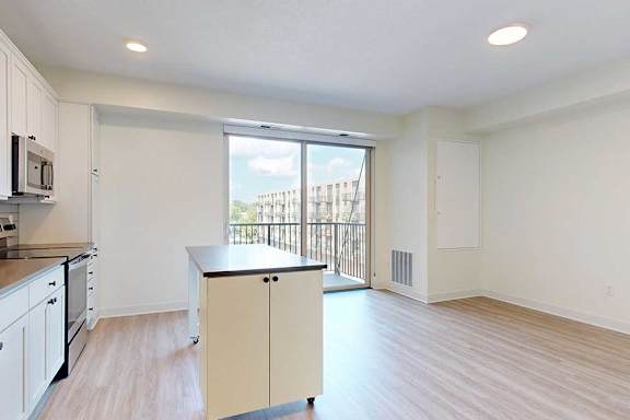 A modern, bright kitchen with white cabinets, stainless steel appliances, and a small island. A large window with a view of a building outside adds natural light.