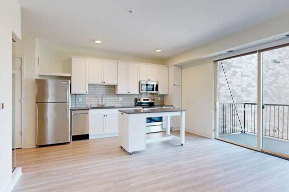Modern kitchen with white cabinets, stainless steel appliances, and a central island on light wood flooring. Large glass doors lead to a balcony.
