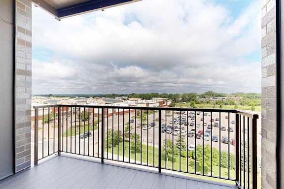 A balcony with a black railing overlooks a parking lot and greenery.