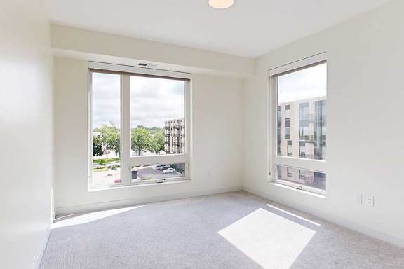 Bright, empty room with large windows, soft ,showcasing natural light and a view of neighboring buildings.