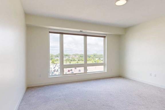 Bright, empty beroom with light-colored walls and carpet, featuring a large window that offers a view of greenery and parking below.