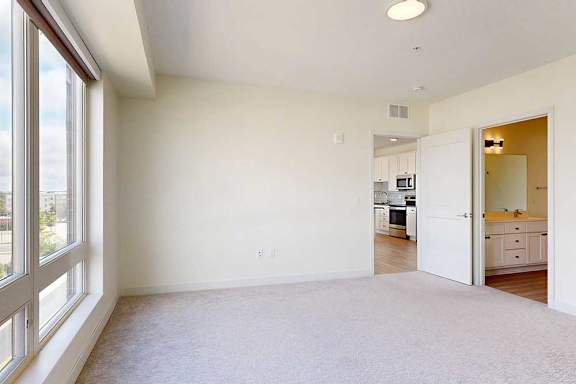 Bright, empty bedroom with beige carpet and a large window. Open doors reveal a modern kitchen and bathroom, creating a clean, inviting atmosphere.
