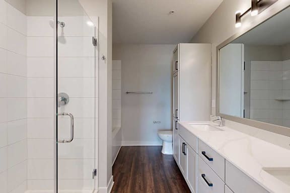 Modern bathroom with a glass shower on the left, wooden floors, white vanity with a large mirror, and a toilet in the background.