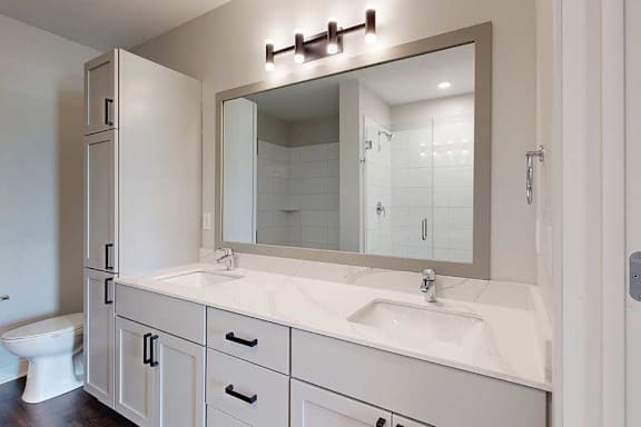 Modern bathroom featuring a dual-sink vanity with white cabinets and black handles, large mirror, overhead lights, and visible toilet and shower.