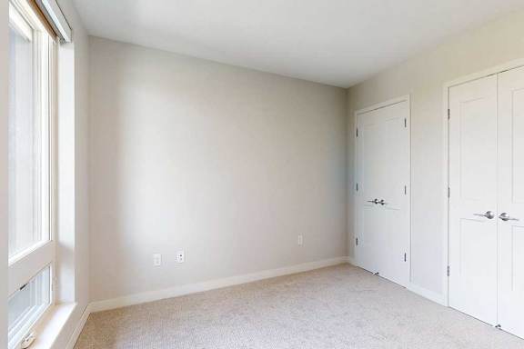 Bright, empty bedroom with beige walls and carpet, large window on the left, and double white closet doors on the right.