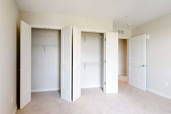 Empty room with light beige walls and carpet, a closet with two open white double-door with shelving.