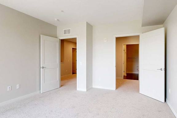 A bright, empty room with light gray walls and beige carpet has two open white doors leading to a hallway and closet.