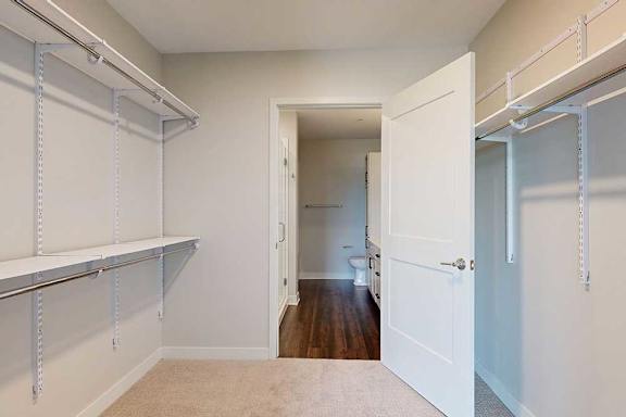 Spacious walk-in closet with beige walls, white shelving on both sides, and an open door leading to a bathroom with wooden flooring.