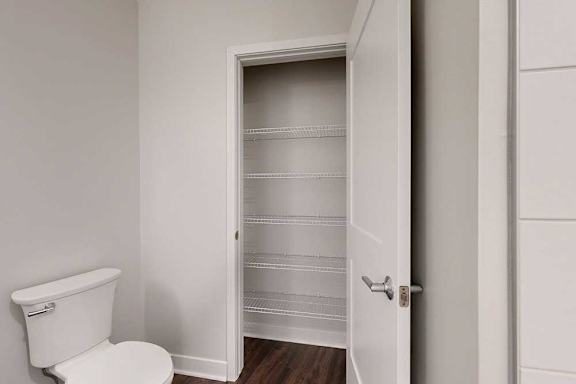Bathroom with white toilet on the left, adjacent to an open door revealing a pantry with empty wire shelves. Walls are light gray, floor is dark wood.