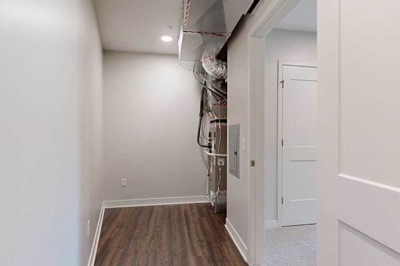 Narrow storage space with hardwood floor and white walls, leading to a utility area with exposed ductwork and piping. A door to the right opens to a carpeted room.