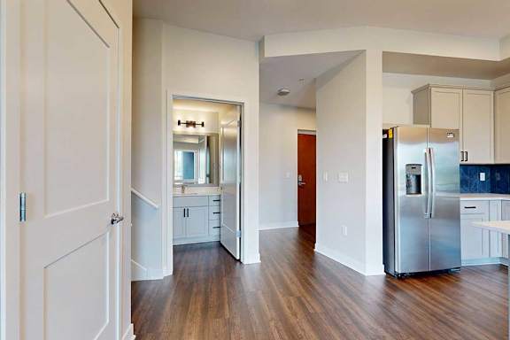 A modern kitchen area features sleek hardwood floors, white cabinetry, and a stainless steel refrigerator. A doorway opens to a bright bathroom.