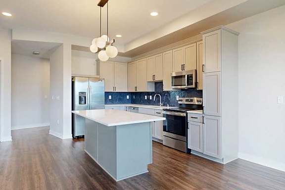 Modern kitchen with white cabinets and a blue backsplash, featuring stainless steel appliances. A central island with a white countertop is illuminated by a stylish pendant light, and dark wooden floors add warmth.