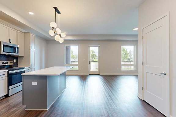 Modern open-concept kitchen and living room with dark wood floors, white cabinets, a gray island, globe pendant light, and large windows offering natural light.