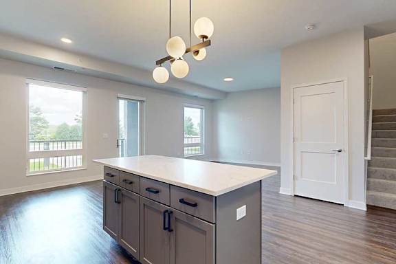 Modern kitchen interior featuring a large island with a white countertop, pendant lights, hardwood flooring, and bright natural light from windows.