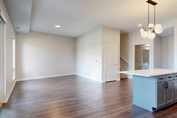 Modern kitchen and living area in neutral tones. Wood flooring, white walls, pendant light fixture, and open island countertop create a spacious feel.