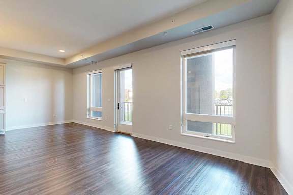 A spacious, modern living room with dark hardwood floors and light walls. Three large windows and a glass door let in natural light, creating an open, airy feel.