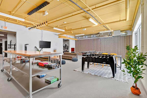 Spacious game room with yellow ceiling, industrial lighting, and board games neatly stacked on a wheeled metal shelf. Bright and inviting atmosphere.