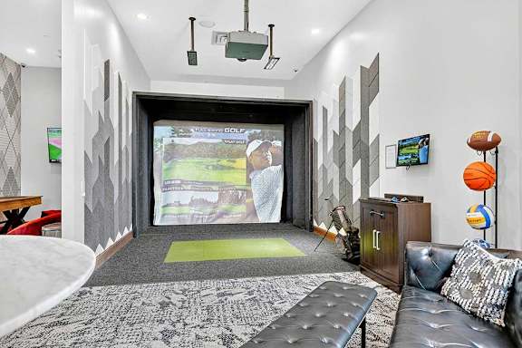 Modern entertainment room with a golf simulator, projector screen, golf clubs, and a cozy seating area. Sports equipment displayed on the wall adds a playful touch.
