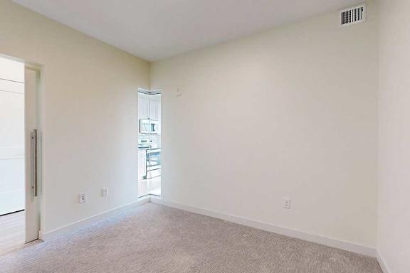 An empty room with beige walls and carpet leads to a bright kitchen visible through a doorway on the left.